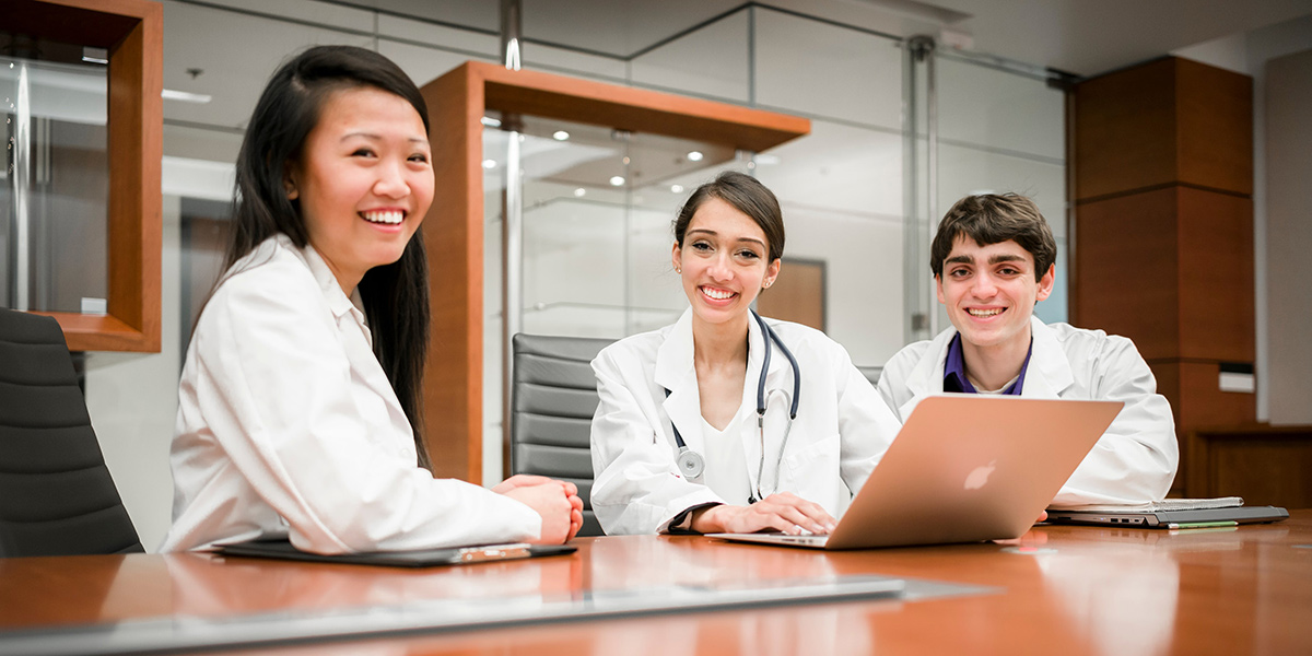 Three Students in White Coats Smiling at a Table