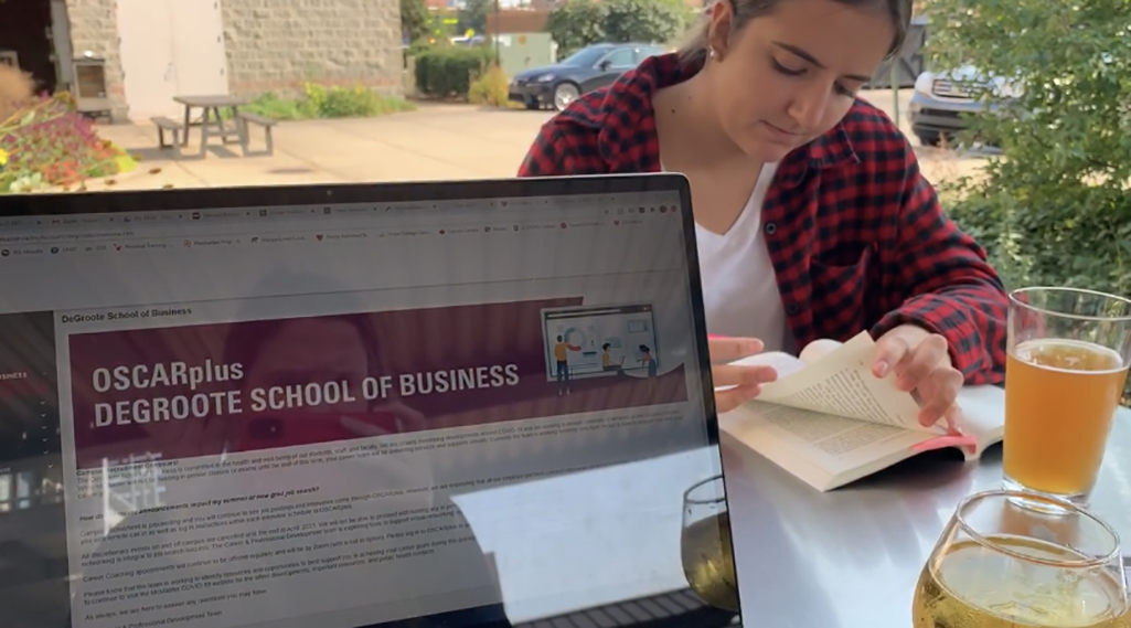 A laptop screen open to the OSCARplus page for the DeGroote School of Business, with a women reading a book in the background