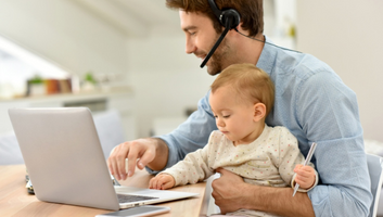 Father working from home on a laptop while wearing a headset and holding his child.