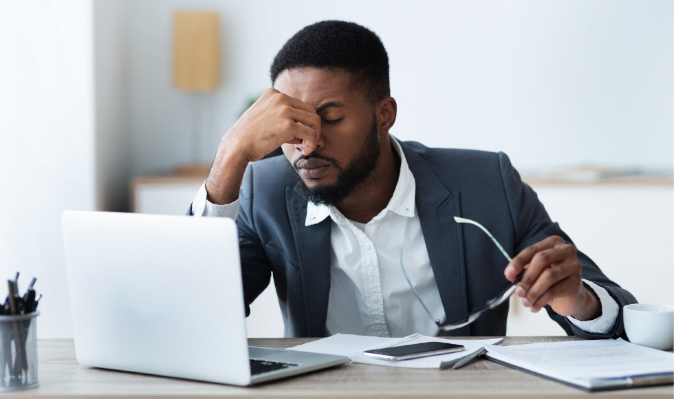 Black male professional in a suit at a desk with laptop, phone, and documents in front of him, holding the bridge of his nose in one hand and a pair of glasses in the other