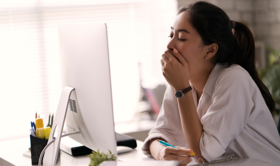 Asian woman with a ponytail at a desk, looking at a computer screen and holding her mouth as she yawns.