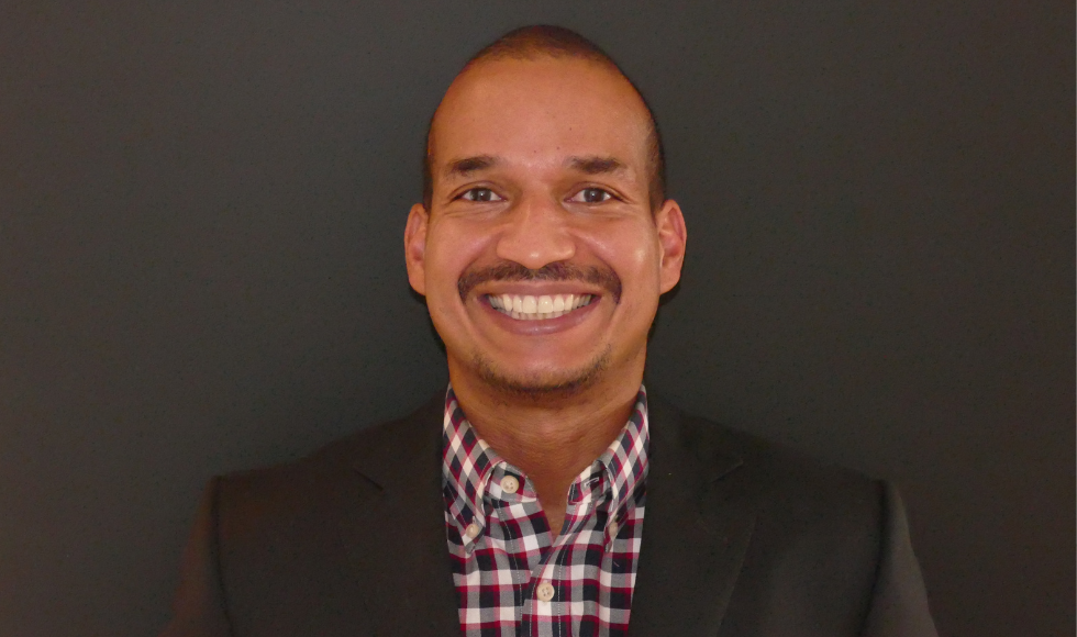 Photo of a man, Earl Davis, C.Dir., facing the camera smiling wearing a black suit and a red white and black checkered shirt, on a dark grey background