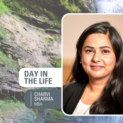 Female student, Charvi Sharma, with long brown hair and brown eyes looking into the camera with a slight smile on a background of nature, rocks, and a waterfall. Includes text "Day in the Life - Charvi Sharma, MBA"