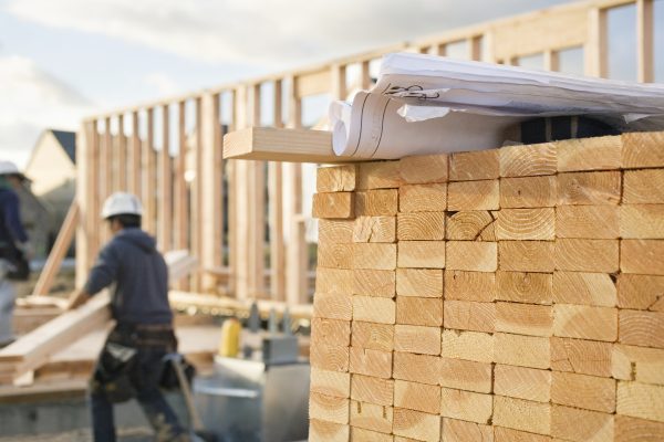 A closeup of stacks of 2x4 boards at a construction site, with a roll of blueprints sitting on top. Two construction workers and a building frame can be seen in the background. Horizontal shot.
