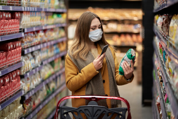 A young woman in a medical mask with a smartphone in her hand scans the grocery store product, contemplating increased food prices.